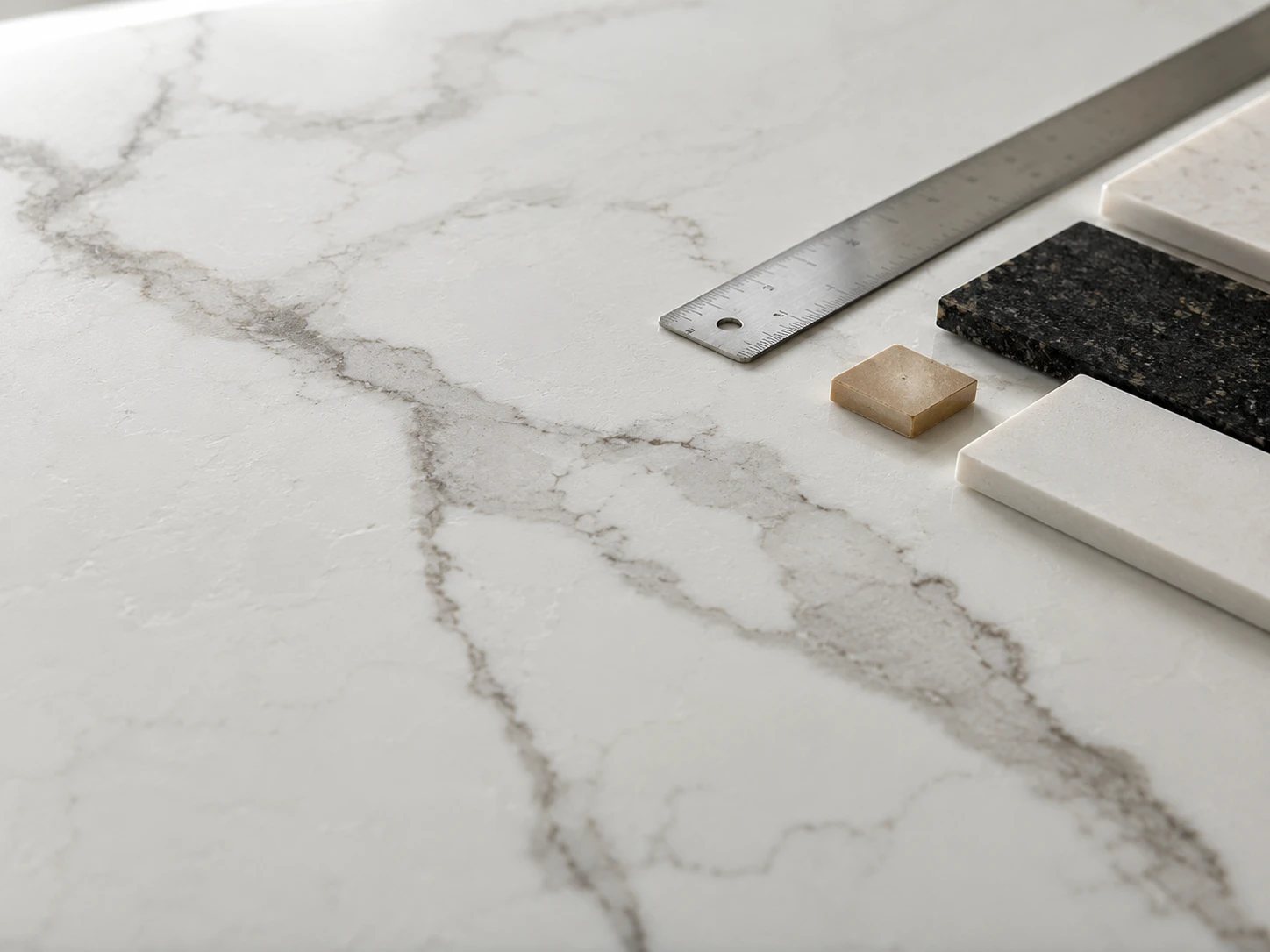 A close view of a printed engineered quartz slab shows controlled marble-like veining, a polished finish, and fine surface texture beside a metal ruler and small color sample chips. Diffused daylight falls evenly across the slab from an overhead skylight. The camera is positioned at a shallow angle on a 50mm lens feel, with the veining line leading diagonally through the frame.