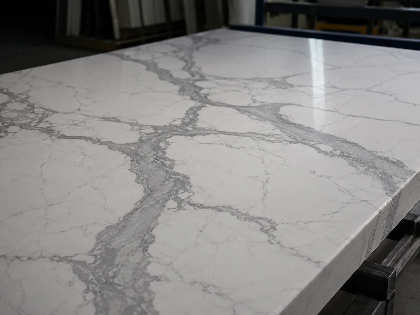 A close-up view of a polished Calacatta-style quartz surface on an inspection table, showing grey veining, resin depth, and a smooth reflective finish. Cool side lighting skims across the slab to reveal texture, flatness, and minor surface reflections. The composition is tight and centered, shot with a natural focal length so the veining becomes the main visual decision point.
