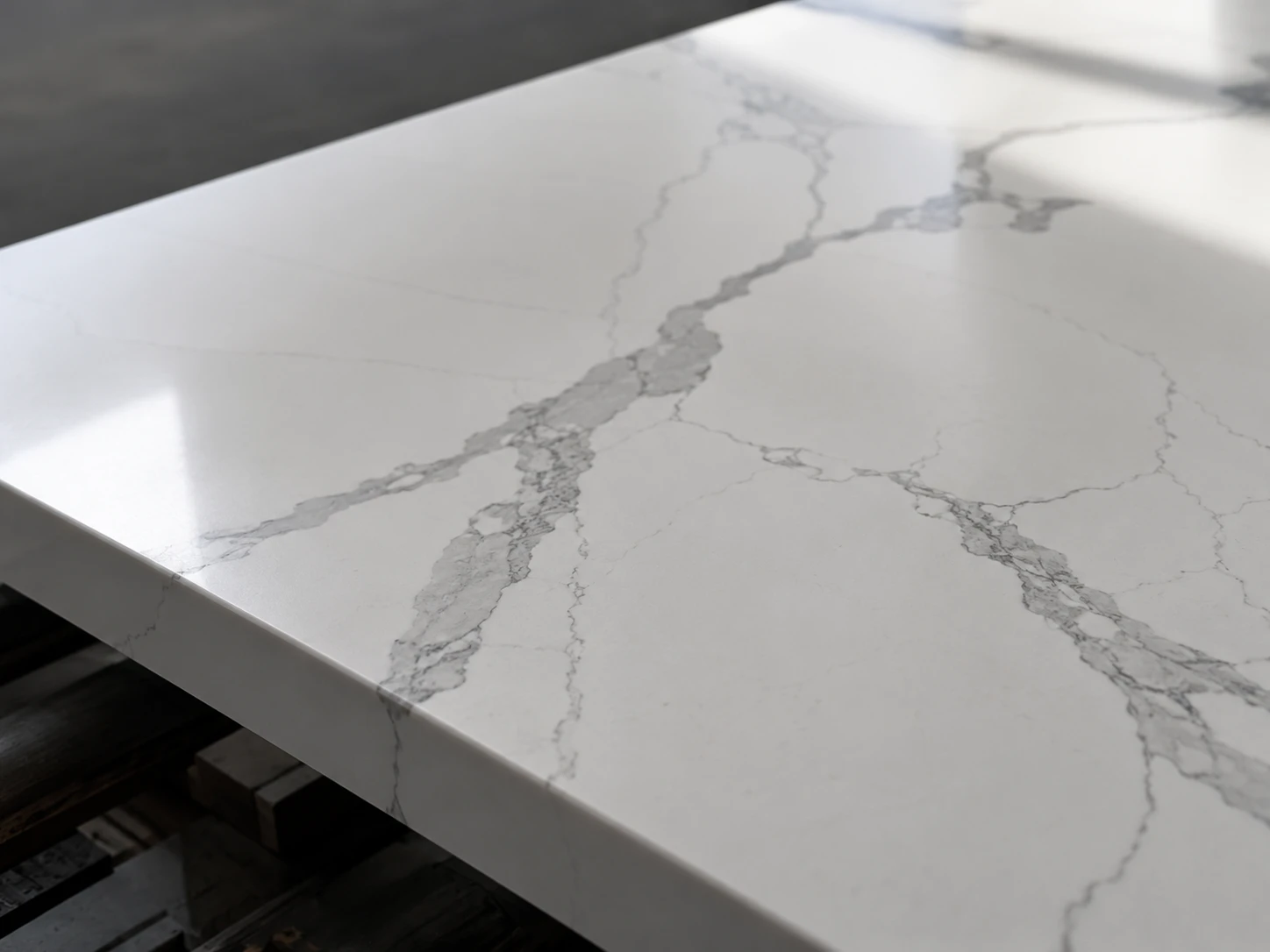 A close-up view of a polished white quartz slab with soft grey veining laid flat on an inspection table. Cool side lighting skims across the surface, showing resin clarity, pattern movement, and a clean reflective finish. Shot on a 50mm lens from a low diagonal angle, with the veining placed along the rule of thirds.