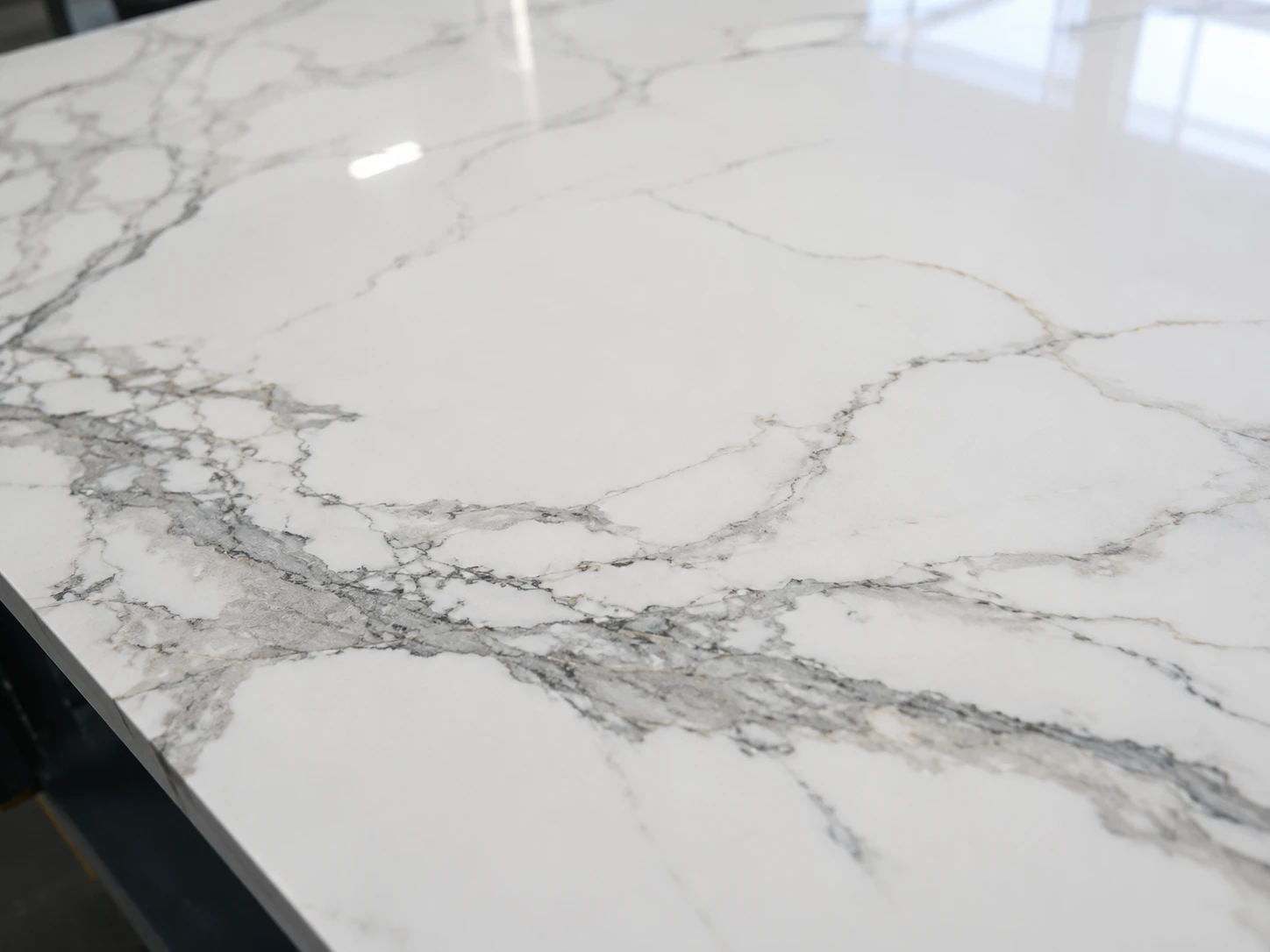 A close view of a polished Calacatta-style quartz slab surface laid flat on an inspection table, with grey veining crossing a clean white background. Cool overhead factory lighting and side daylight reveal resin sheen, fine particulate detail, and reflection consistency. The camera looks down from a shallow diagonal angle, framing the veining across the lower third with quiet negative space.