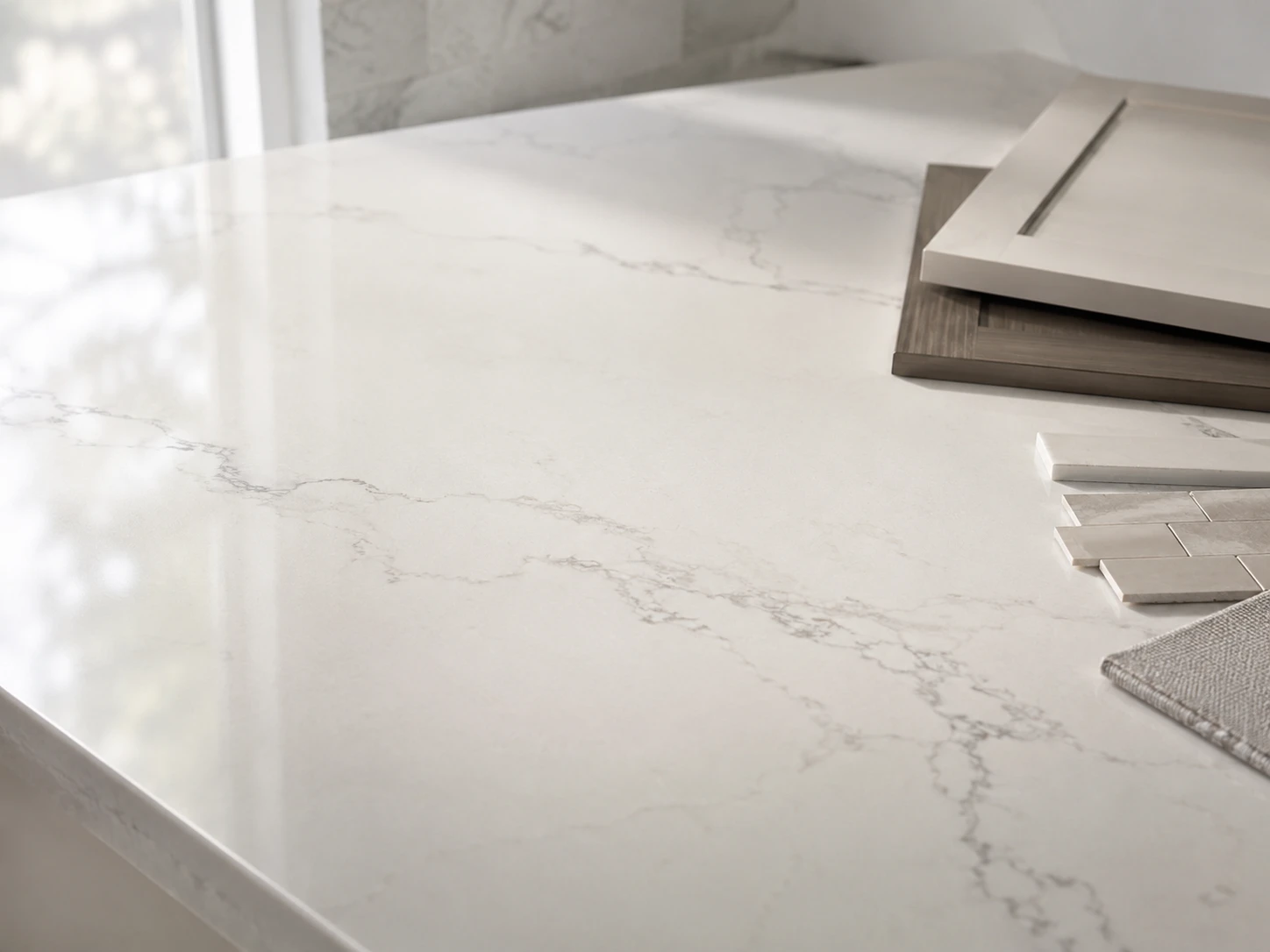 A close-up of a light Calacatta-style quartz surface on a worktable, showing subtle grey veining, polished reflection, and a small set of neutral cabinet and backsplash samples nearby. Soft morning window light skims across the surface from the left, photographed from a slightly elevated 50mm angle with the veining placed along the rule of thirds.